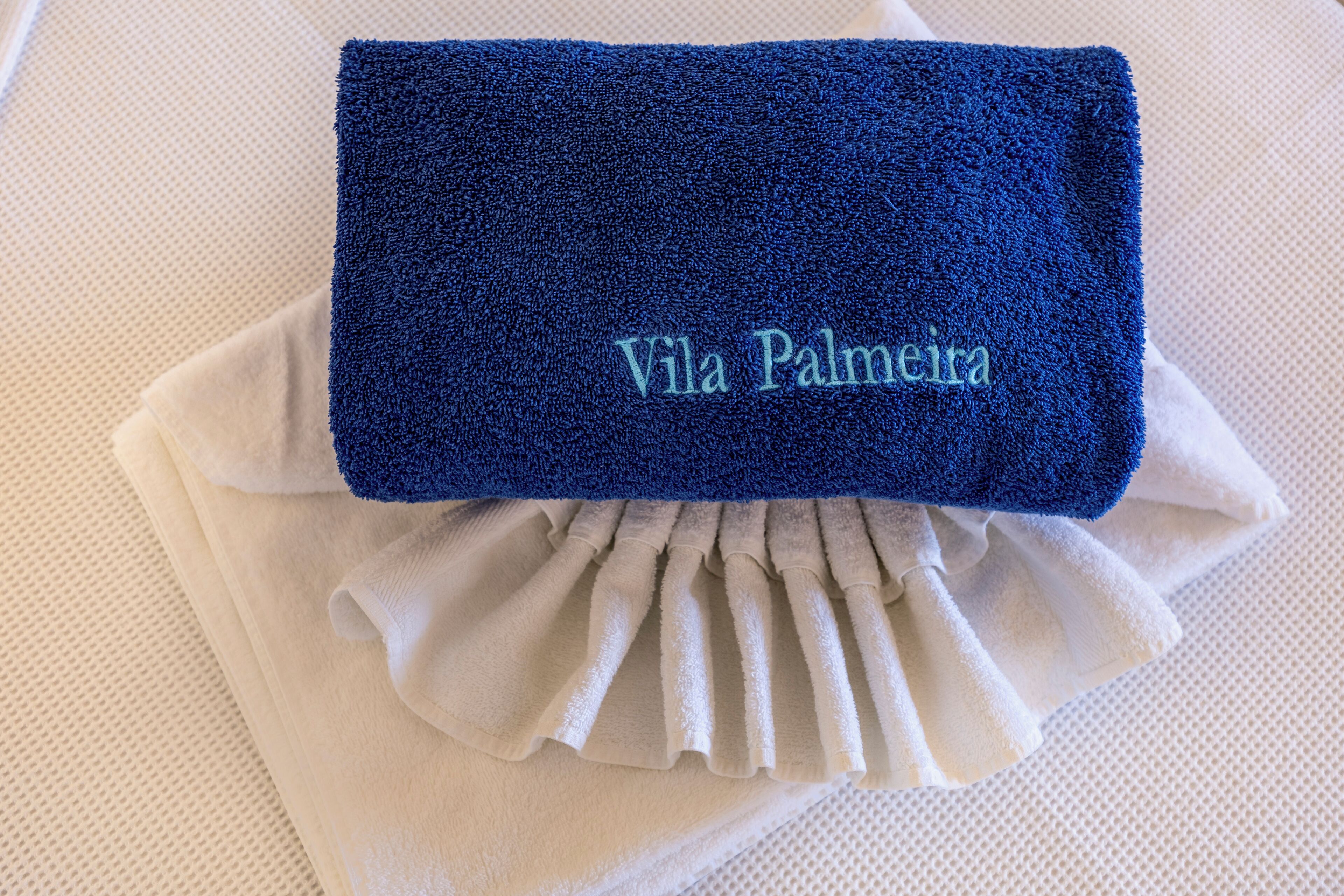 Une serviette bleue, soigneusement pliée et ornée d’une broderie « Vila Palmeira », est posée sur une surface blanche à texture.