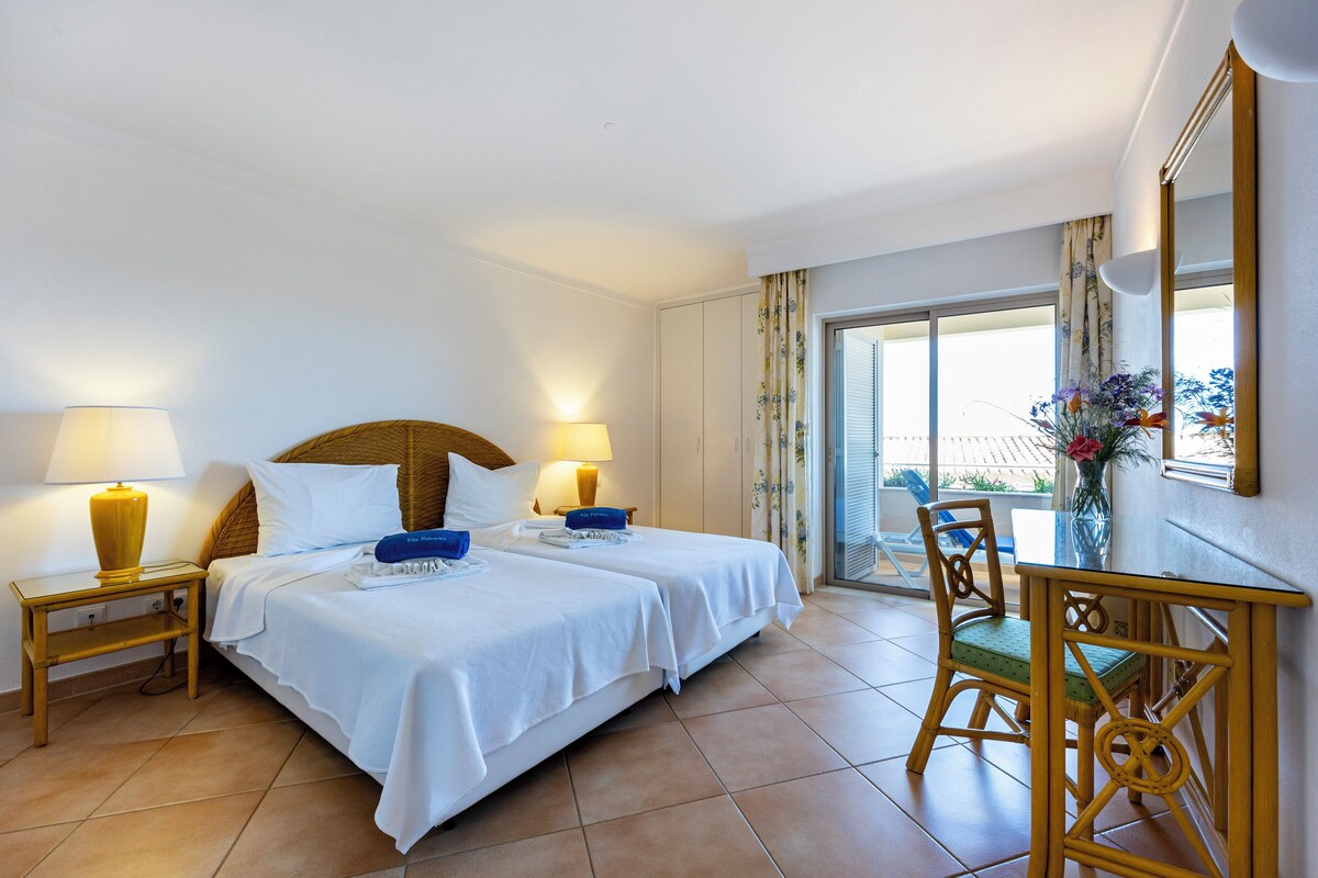 A hotel room with two beds, a dining table, chairs, lamps, and a large window with curtains.