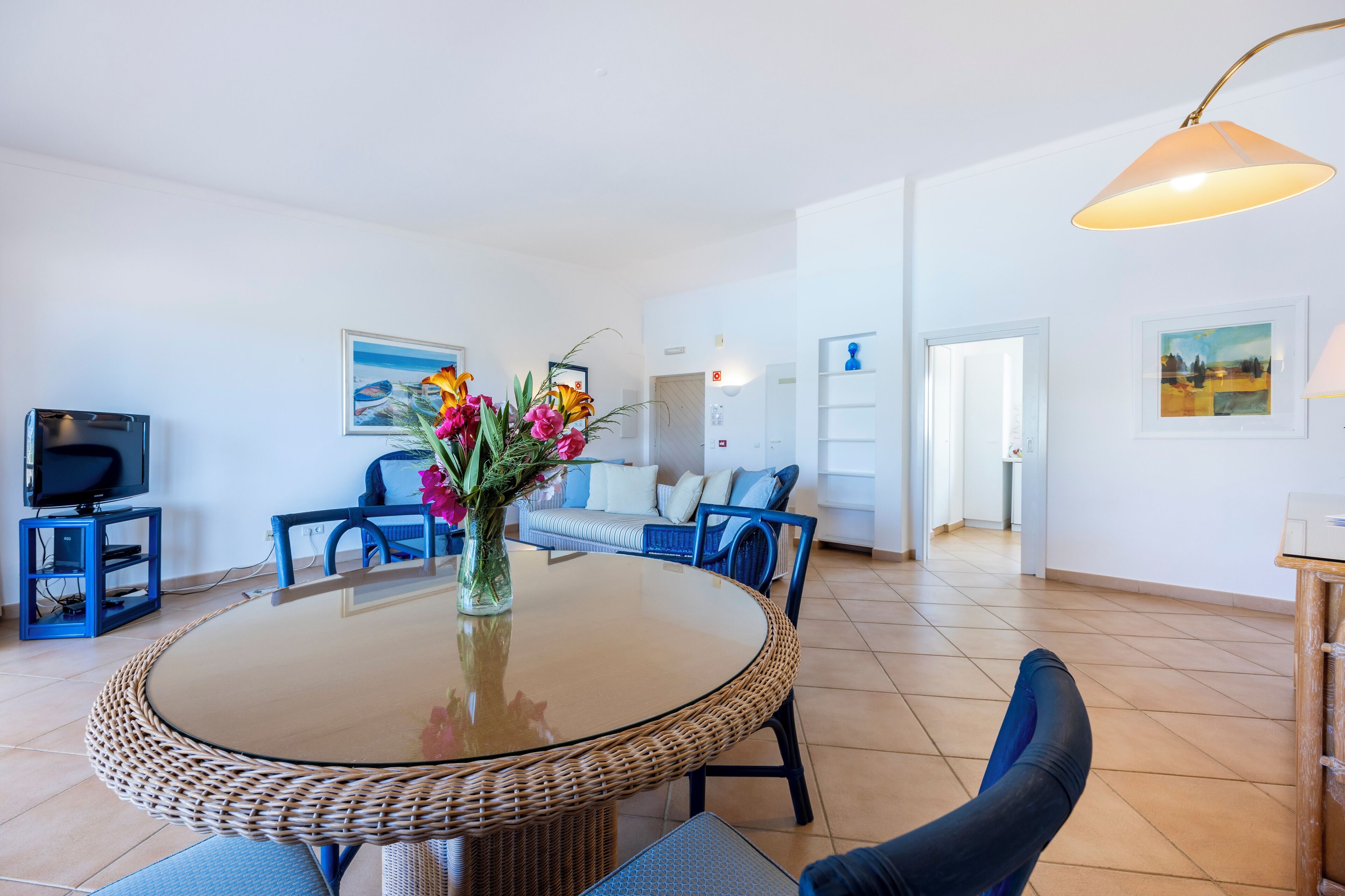 A modern living room with a round glass-top table, blue chairs, a sofa, a TV on a stand, and a vase with flowers.