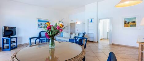 A modern living room with a round glass-top table, blue chairs, a sofa, a TV on a stand, and a vase with flowers.