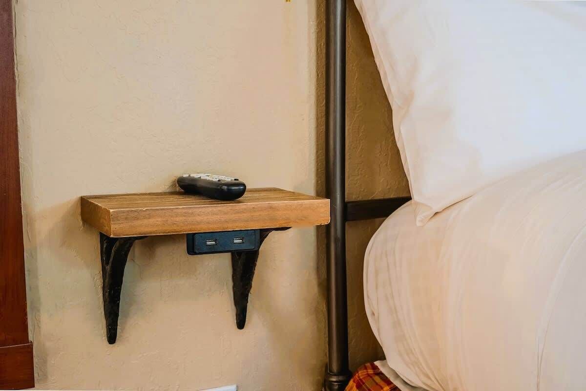 Uma mesa de cabeceira com um controle remoto, uma prateleira fixada na parede e uma cama com roupa de cama branca.