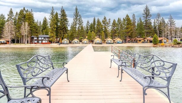 Une jetée en bois menant à un lac, avec des bancs, entourée d’arbres et de cabanes.