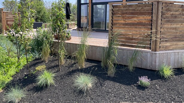Una casa moderna con terraza de madera, ventanas amplias y jardín con diversas plantas.