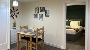 Un dormitorio con cama, mesita de noche, una silla y una pequeña mesa con lámpara. Se observan adornos en la pared y una puerta que da a otra habitación.