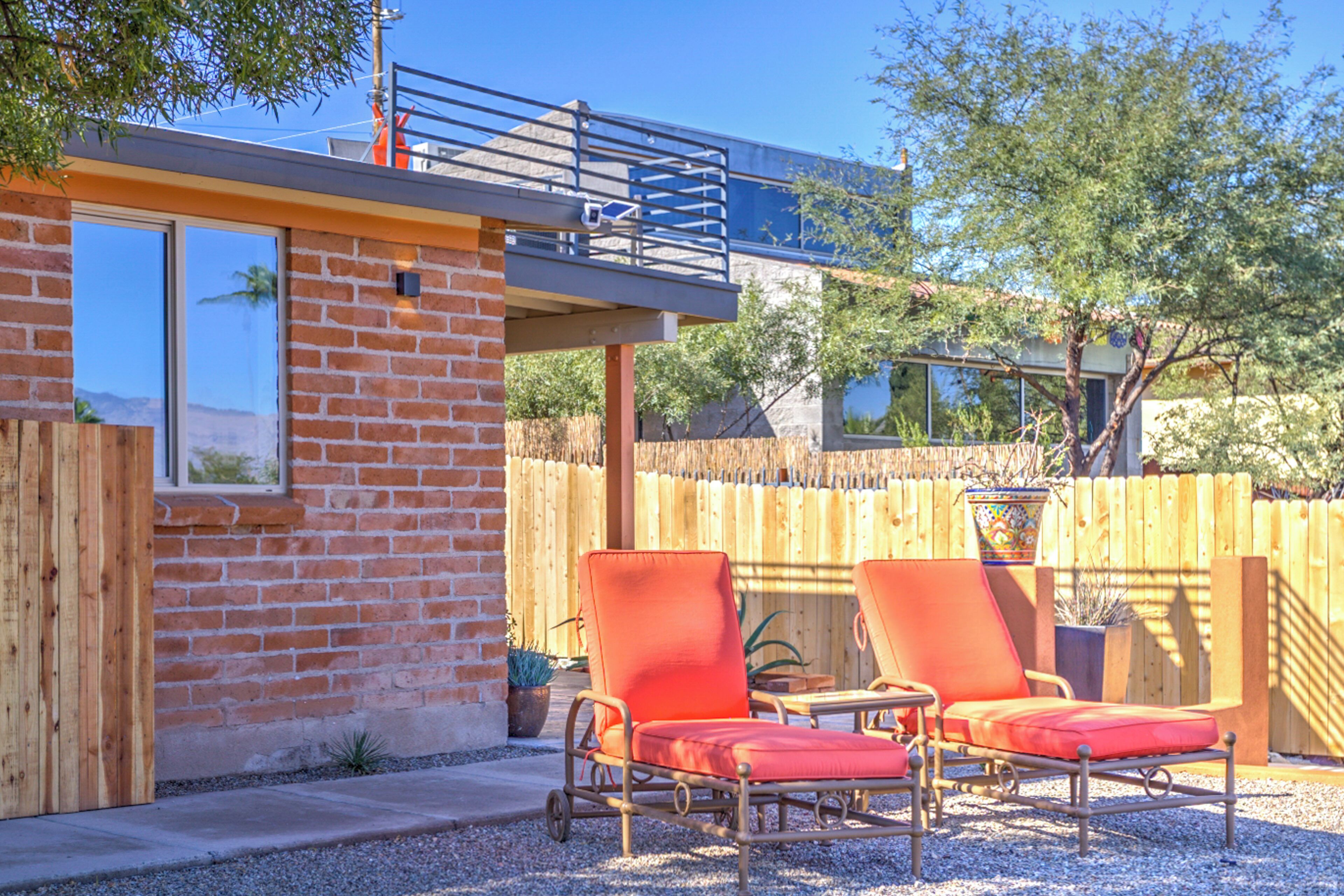 A patio area with two orange chairs and a small table, a brick building with a wooden deck, and a wooden fence.