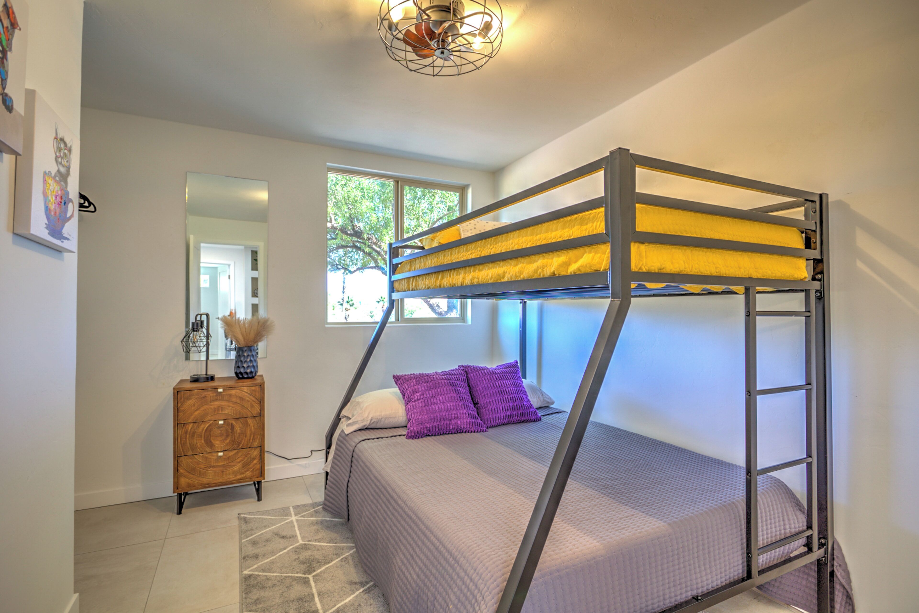 Un lit superposé avec une structure en métal et une literie jaune, une commode en bois, un miroir et un tapis à motifs géométriques.