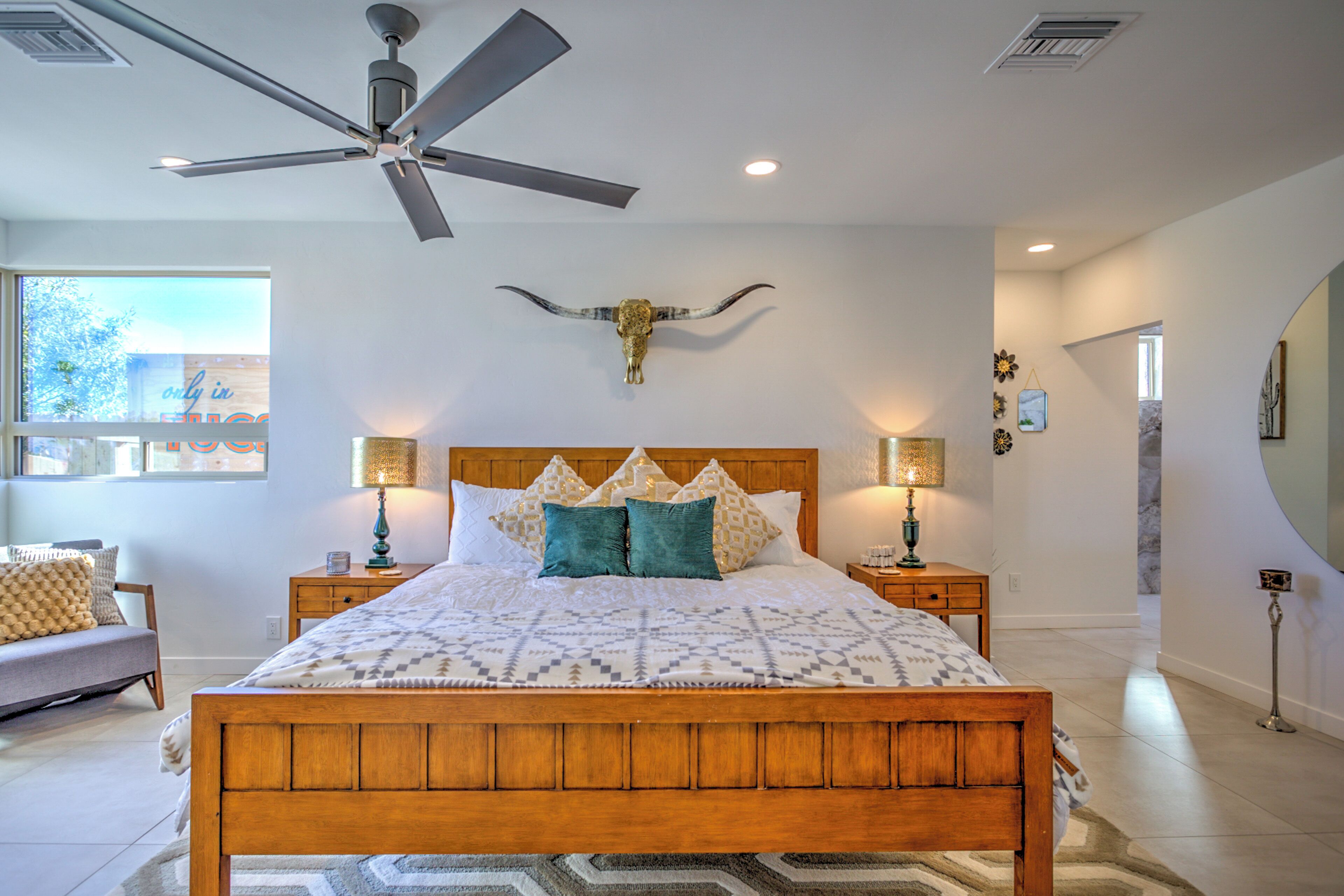 A bedroom with a large bed, wooden headboard, two bedside tables with lamps, a ceiling fan, and a decorative bull skull on the wall.
