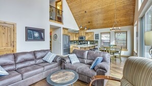 A spacious living area with a large leather sofa, a round coffee table, and a striped armchair. A kitchen with wooden cabinets and a dining area with a table and chairs are visible in the background. - Angel Fire Cabin w/ Deck: 3 Mi to Ski Lifts! (Angel Fire)