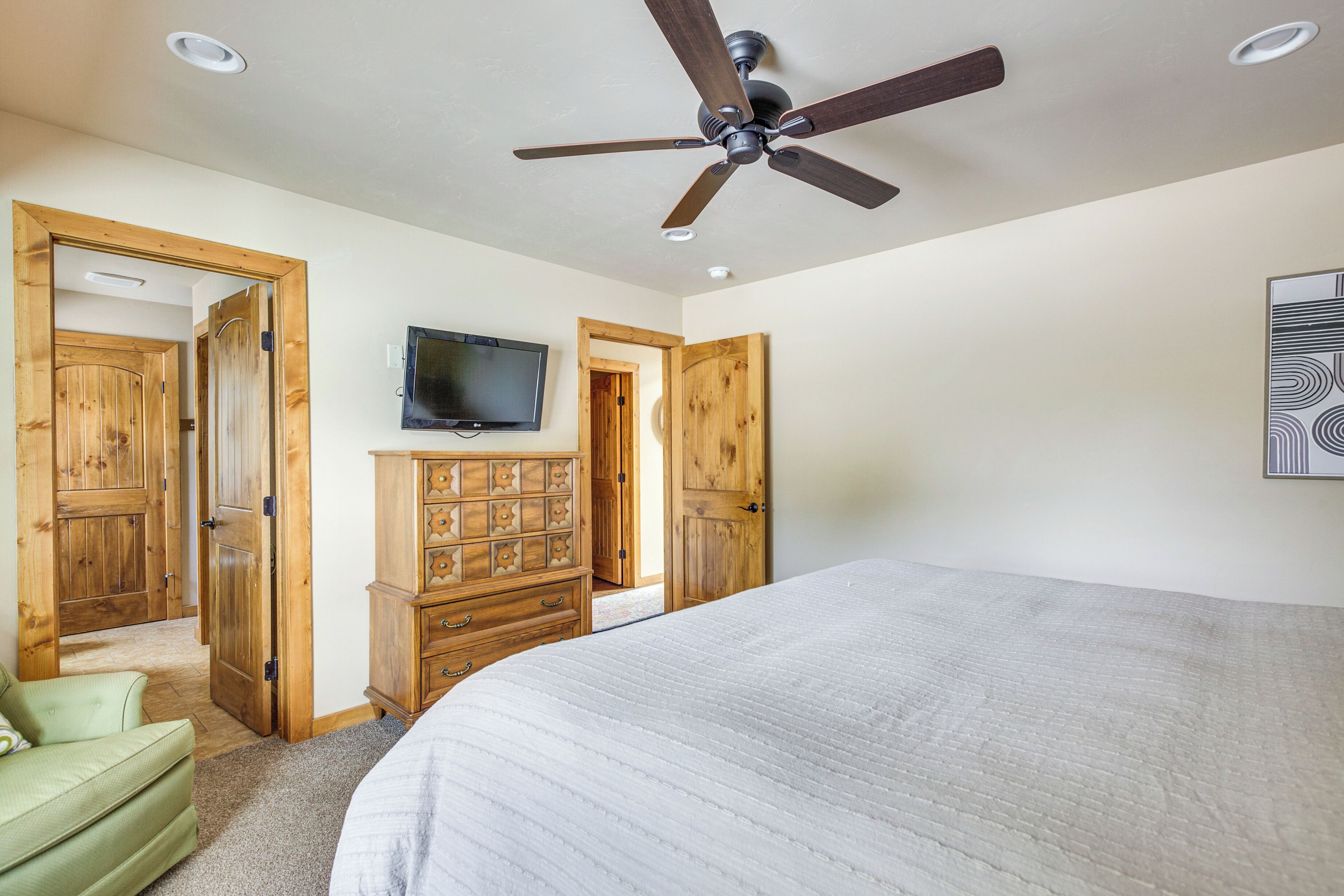A bedroom with a large bed, wooden wardrobe, a ceiling fan, and a TV mounted on the wall.