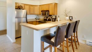 A modern kitchen with wooden cabinets, a stainless steel refrigerator, a microwave, and a bar area with stools.