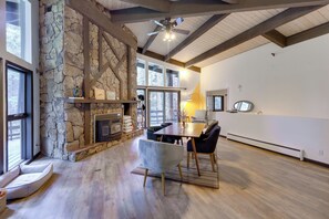 A spacious living area with a stone fireplace, wooden beams, a dining table with chairs, and large windows offering views of the outdoors. - Mid-century Cabin: Creekside, Easy Access to I-70 (Dumont)