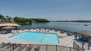 Ein Schwimmbecken mit Liegestühlen und Sonnenschirmen, umzäumt von einem Zaun, mit Blick auf einen See und Gebäude im Hintergrund.