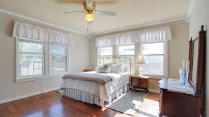 Une chambre à coucher comprenant un lit, des tables de chevet, une commode, un meuble à tiroirs, un miroir, un ventilateur de plafond et de grandes fenêtres.