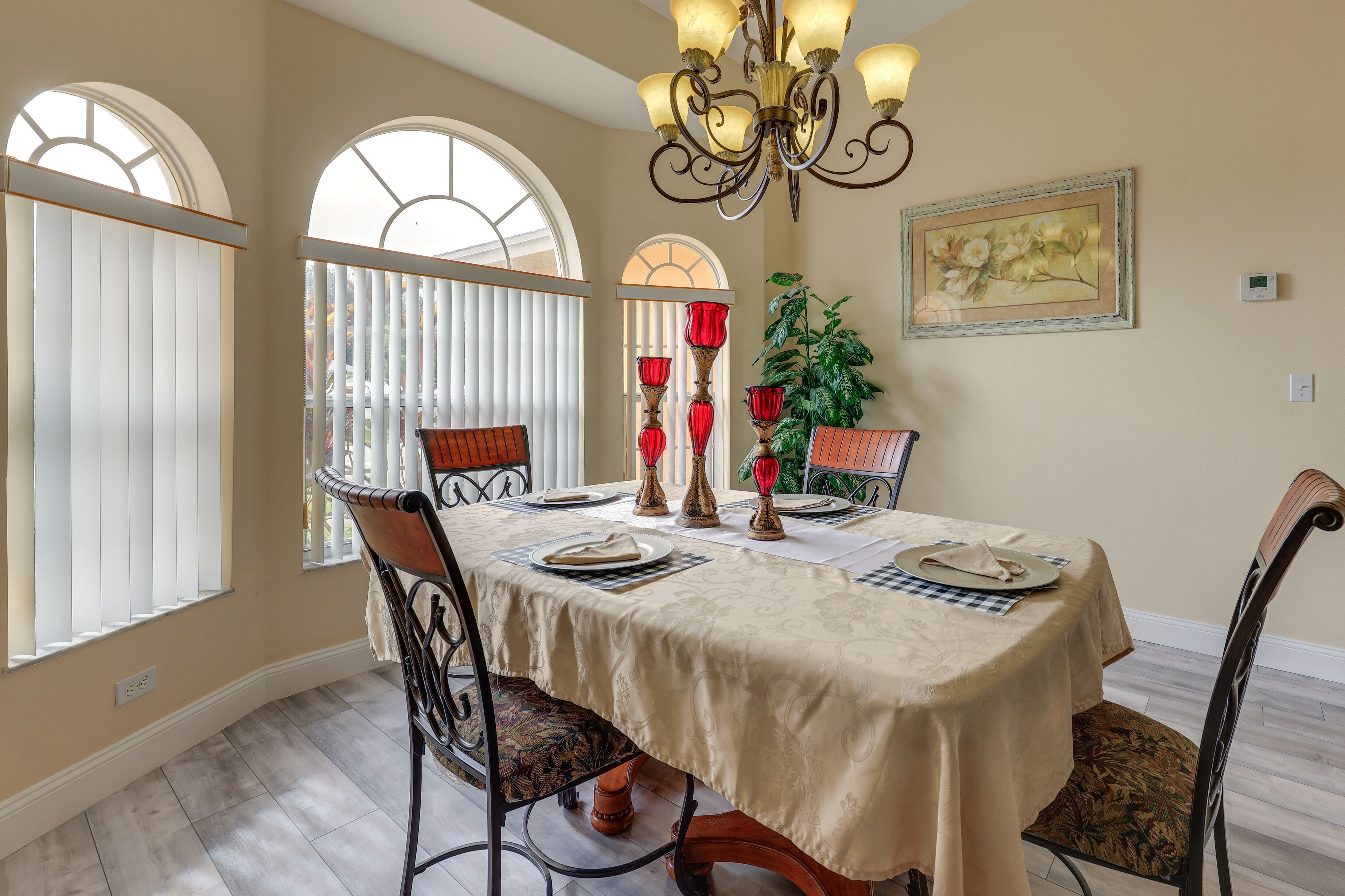 A dining room with a table set for four, featuring a chandelier, red candle holders, and a potted plant.