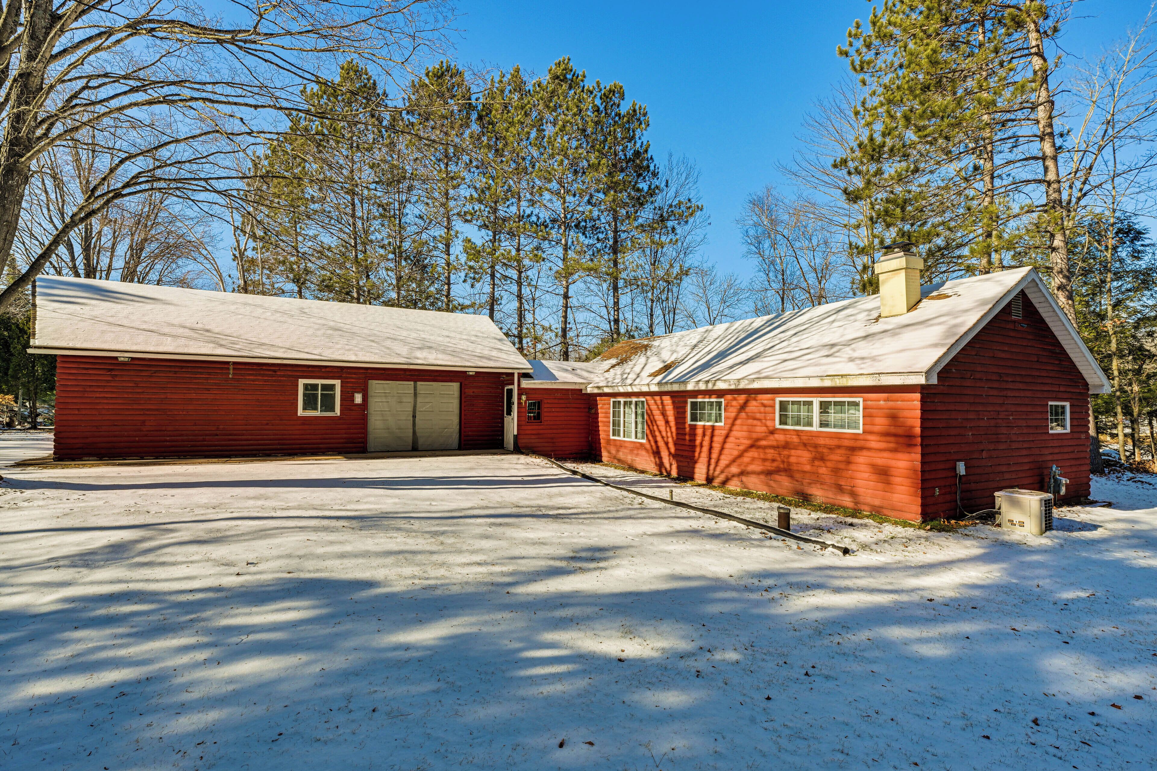 A two-story red wooden cabin with a garage, surrounded by trees and snow.