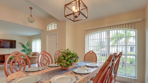 A dining area with a long table set for six, surrounded by wooden chairs, a decorative centerpiece, and a large window with blinds. - Pet-friendly Port St Lucie Home ~ 5 Mi to Marina! (Port Saint Lucie)