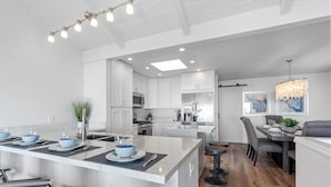 A modern kitchen with white cabinetry, a central island, and a dining area with a wooden floor. - Cardiff Panoramic Paradise - Stunning Ocean Views (Cardiff By The Sea)