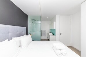 Quarto de hotel moderno com uma cama grande, box de vidro para o chuveiro, banheiro branco e uma porta que dá para outro quarto.