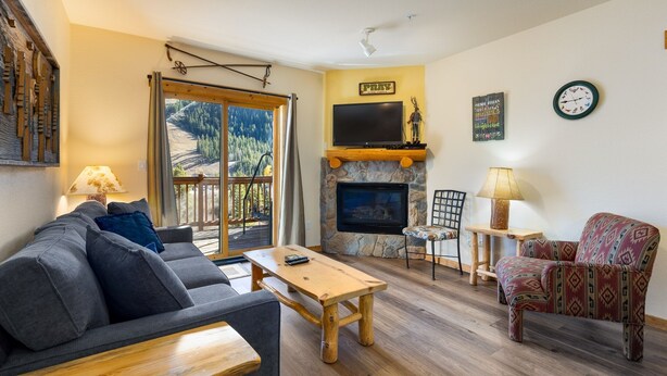 A living room with a sofa, a wooden coffee table, a patterned armchair, a stone fireplace, a TV mounted above the fireplace, and a large window with a view of trees and mountains. - 2656 Tenderfoot Lodge 2 Bedroom Condo by RedAwning (Keystone)