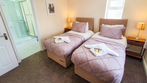 A hotel room with two beds, each with a headboard, a bedside table, and a lamp. There is a door leading to a bathroom with a glass shower enclosure. - Charming 2-bed Apartment in Stroud (Stroud)