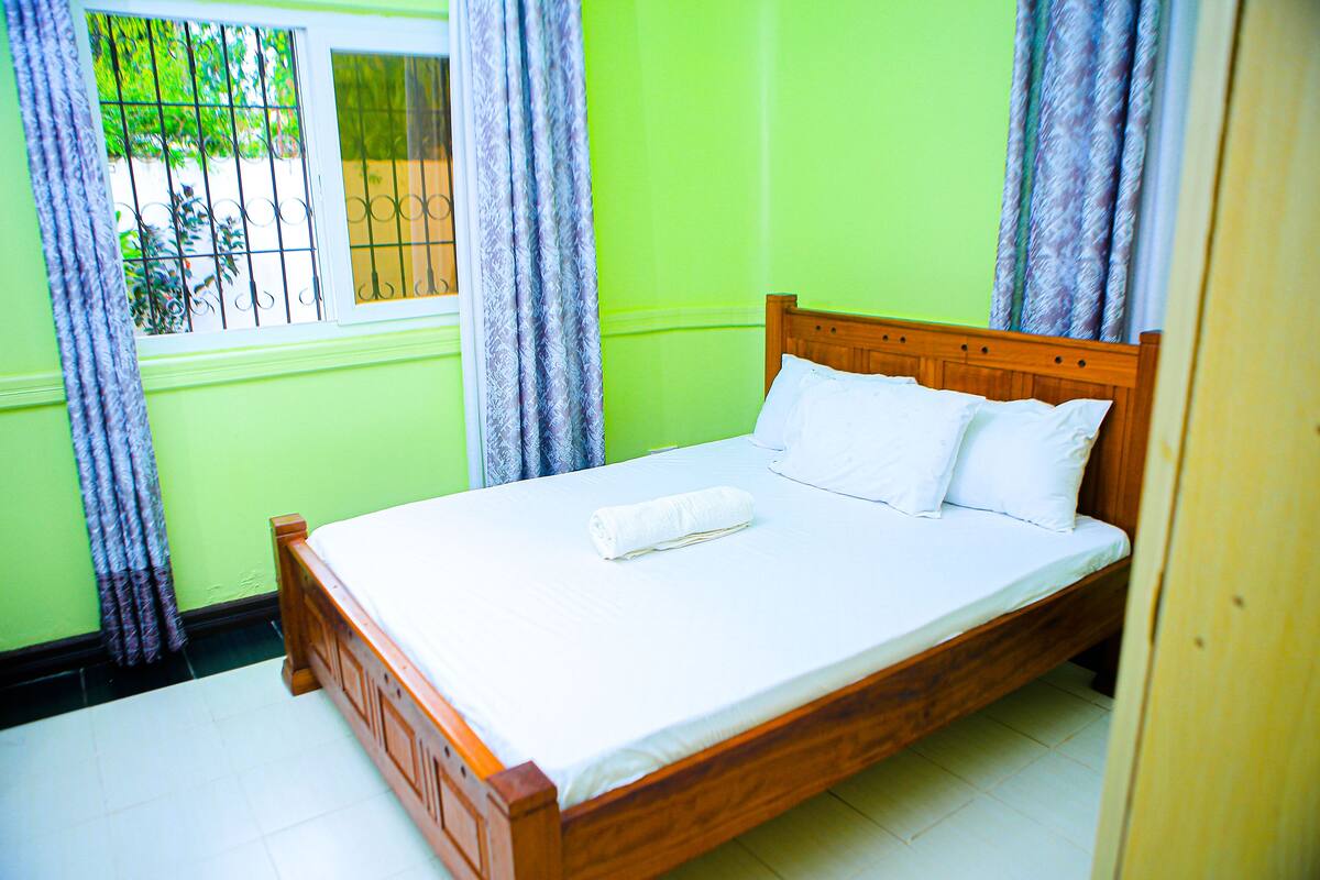 A single-bed room with a wooden headboard, white bedding, and a window with metal bars.