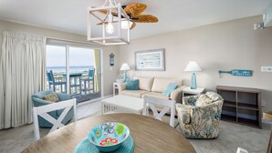 A living room with a view of the beach, a round table with a colorful bowl, and a ceiling fan.