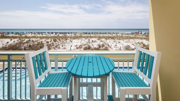 A balcony with two turquoise chairs and a small table, overlooking a beach with sand dunes and the ocean.