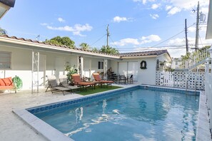 Una piscina con sillones de descanso y una pequeña mesa, rodeada por una cerca blanca y una casa con tejado de tejas.
