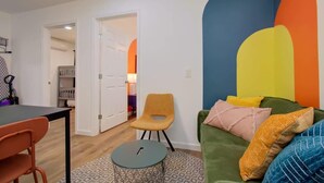 Una sala de estar con un sofá verde, una mesa redonda para café y una silla amarilla. Hay una puerta que da a otra habitación.