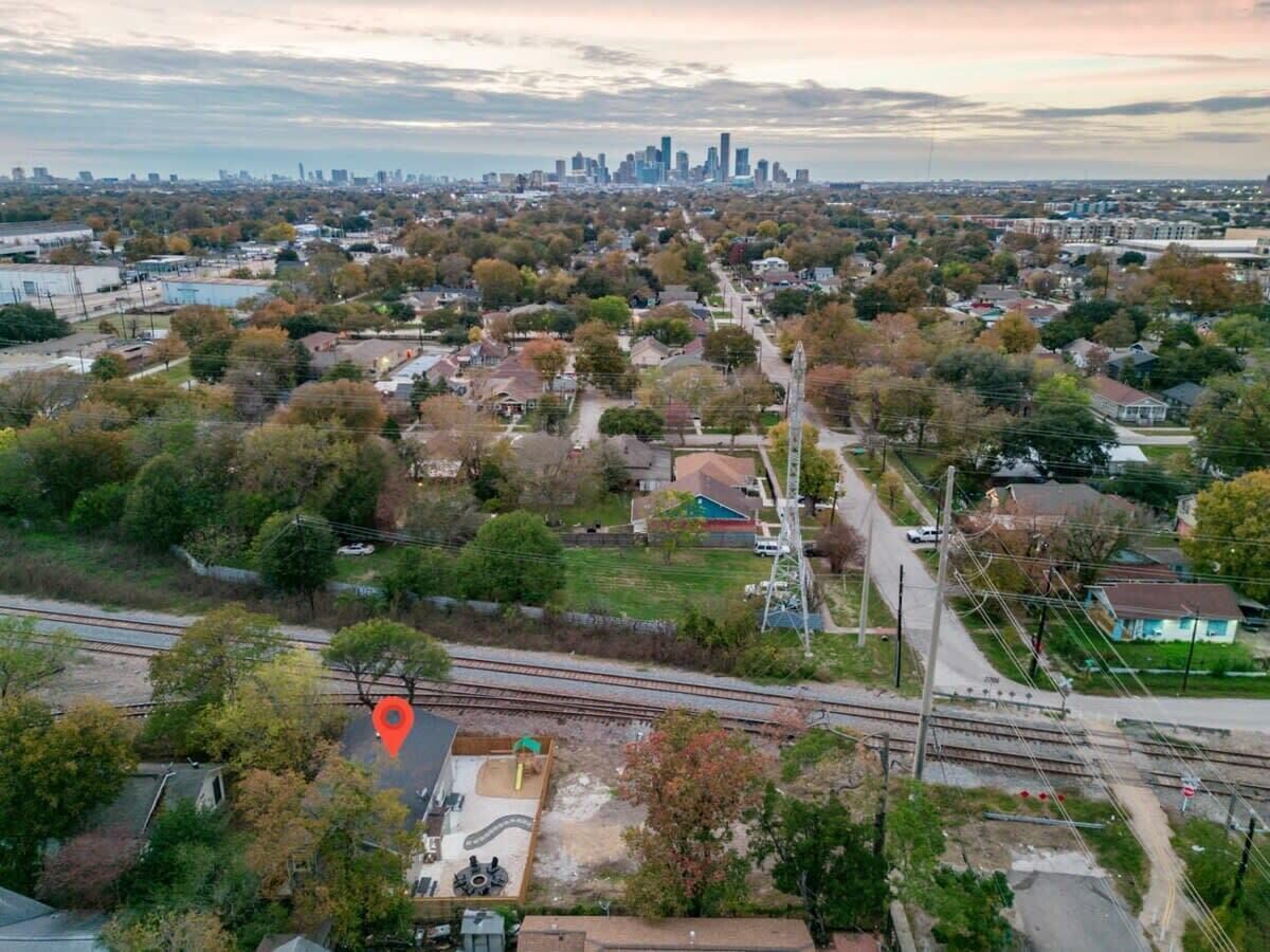 An aerial view of a suburban area with a railway track, residential houses, and a distant city skyline.