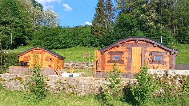 Dos cabañas de madera rústicas con techos a dos aguas, rodeadas de exuberante vegetación y un muro de piedra.