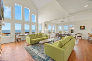 A spacious living room with large windows, a green sofa, a dining area, and a view of the sea. - Soundfront Home w/ Pool, Hot Tub, Elevator, Private Windsports Access (Salvo)