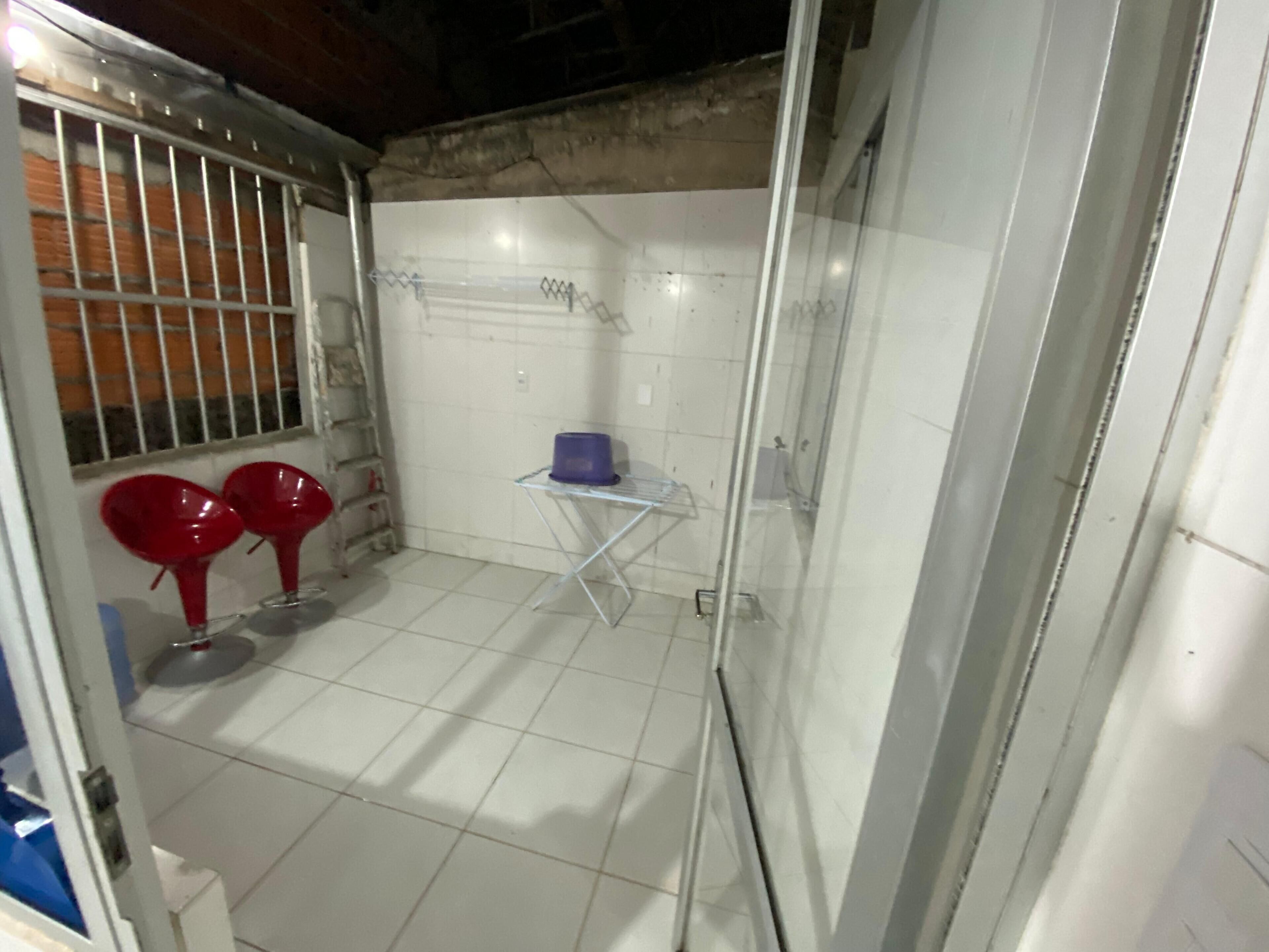 A small, enclosed bathroom with white tiles, a metal door with a window, and a small table with a blue container.