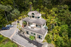 A modern two-story house with a curved roof, surrounded by lush greenery and palm trees. - Bayview Panorama - Paihia Holiday Home (Paihia)