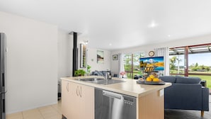 A modern kitchen with a dishwasher, a sink, and a built-in oven. There is a dining area with a blue sofa and a TV. The room has a large window with a view of greenery outside. - Summer Haze - Omaha Holiday Home (Matakana)