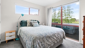 A bedroom with a bed, a nightstand, a desk lamp, and a large window with a view of trees and a red structure outside. - Summer Haze - Omaha Holiday Home (Matakana)
