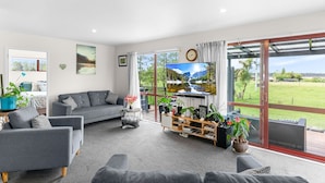 A spacious living room with a grey sofa set, a coffee table, and a TV on a stand. There are potted plants and a bookshelf. The room has large windows with a view of a green landscape. - Summer Haze - Omaha Holiday Home (Matakana)