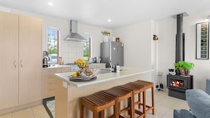 A modern kitchen with a central island, wooden bar stools, a built-in stove, and a seating area with a sofa and a potted plant. - Summer Haze - Omaha Holiday Home (Matakana)
