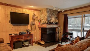 A cozy living room with a stone fireplace, a television mounted on the wall, and a view of a snowy outdoor area through a large window. - Canyons View 7: 2 Bedroom 2 bath vacation condo (Park City)