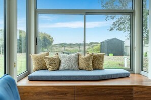 A window seat with a cushioned bench and patterned pillows, overlooking a grassy area with a building in the distance.