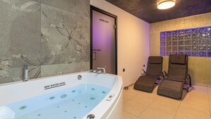 Un área de spa con una bañera grande, dos tumbonas, una pared con estampado floral y una puerta iluminada con luz púrpura.
