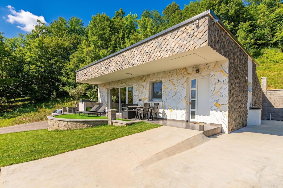 A modern house with a stone exterior, a patio with outdoor furniture, and a view of a lush green forest.