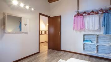 Ein Schlafzimmer mit Holzboden, einem Bett mit blau-weiß gestreifter Decke, einem Holzschrank, einem Spiegel und einem Kleiderständer mit verschiedenen Kleidungsstücken.