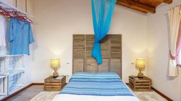 Ein Schlafzimmer mit einem Bett, einem Holzkopfteil, Nachttischlampen, einem hängenden blauen Baldachin und einem weißen Kleiderschrank mit gestreifter Kleidung.