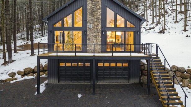 Une cabane moderne dotée d’une cheminée en pierre, de grandes fenêtres et d’une terrasse en bois.