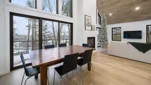 Un salon moderne avec une table à manger en bois, des chaises noires, une cheminée, une grande fenêtre et un téléviseur fixé au mur.