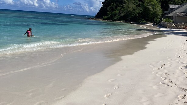 Ein Strand mit klarem türkisblauem Wasser, einem sandigen Ufer und einer bewaldeten Hügelkette im Hintergrund.