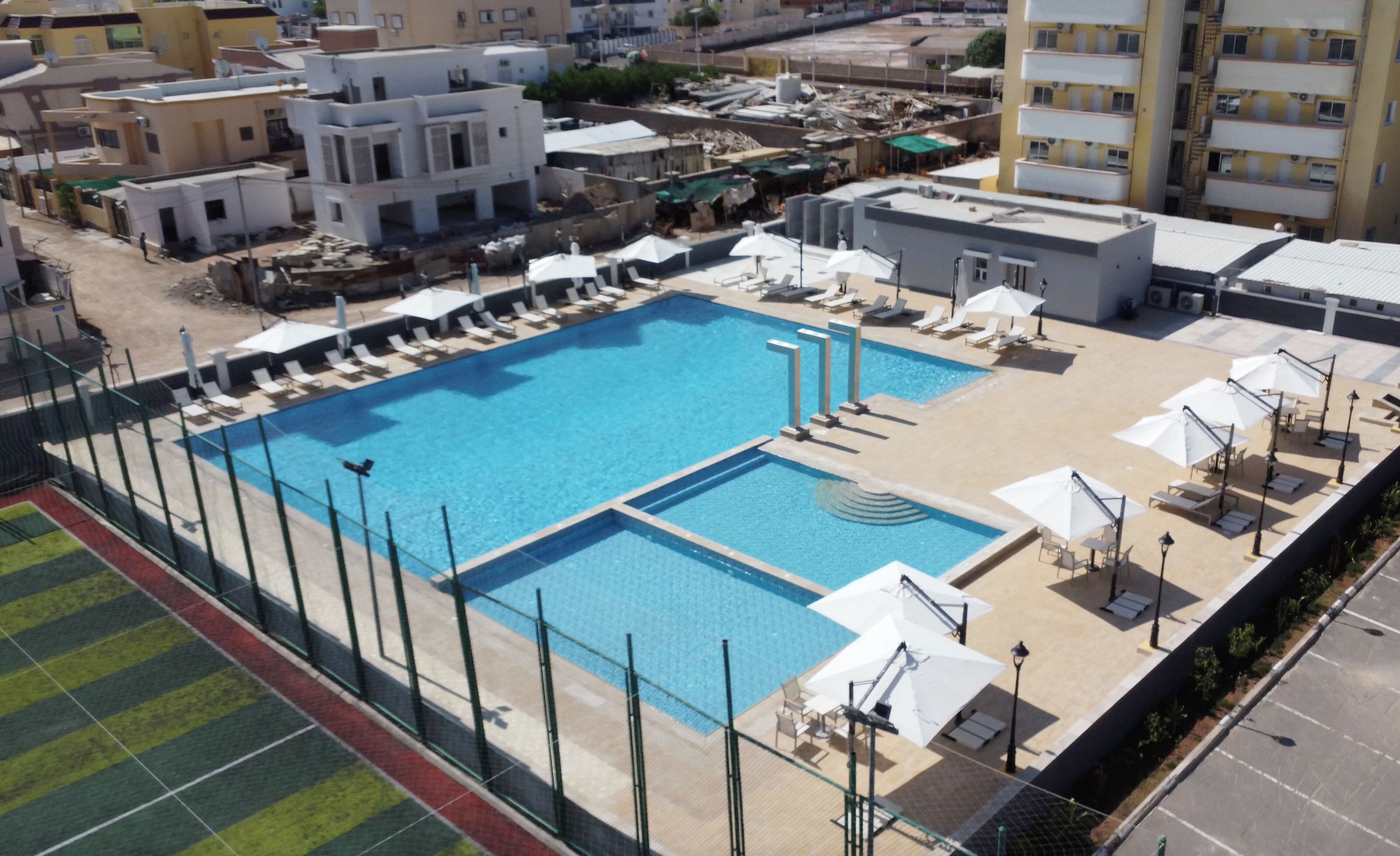 Une grande piscine extérieure dotée de plusieurs couloirs, entourée de chaises longues et de parasols, avec en arrière-plan un ensemble d’immeubles résidentiels.