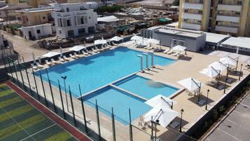 A large outdoor swimming pool with multiple lanes, surrounded by sun loungers and umbrellas, set against a backdrop of residential buildings.