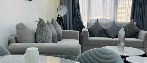 Una sala de estar moderna con un conjunto de sofás, una mesa de comedor con platos y cubiertos, y un ventanal con cortinas.